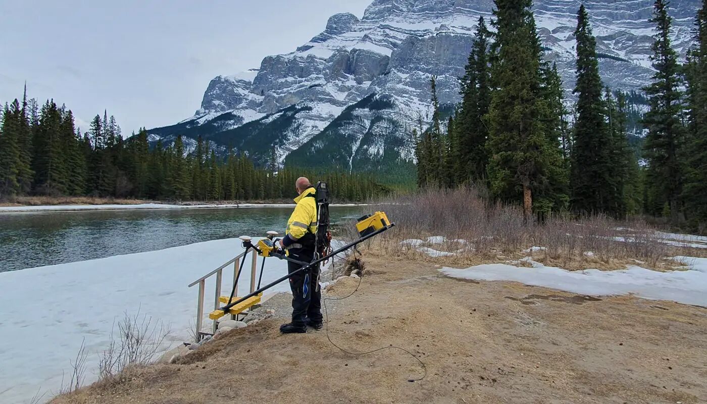 An inspector is standing at the shore of a river in front of the rocky mountains
