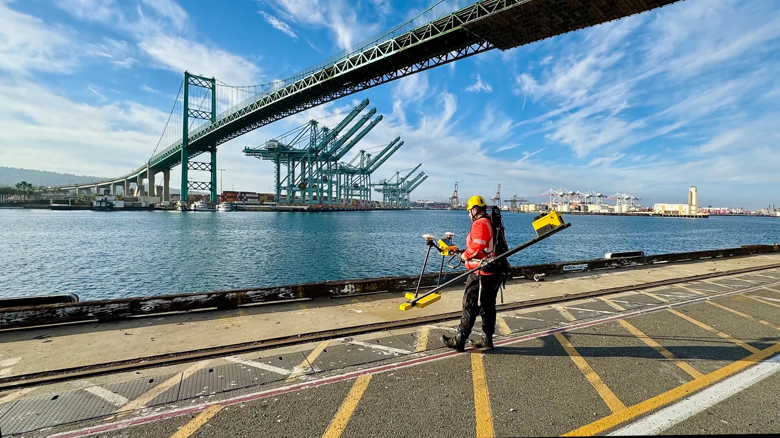 An EMPIT technician in a high-visibility vest and safety helmet walks along the harbor edge while operating the Current Mapper inspection device. Behind him, towering container cranes line the dock beneath a sweeping suspension bridge.