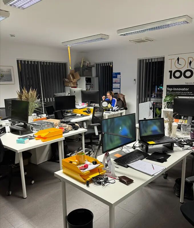 One of the modern open-plan offices in Berlin filled with white desks arranged in clusters, each topped with multiple computer monitors, laptops, and piles of documents.