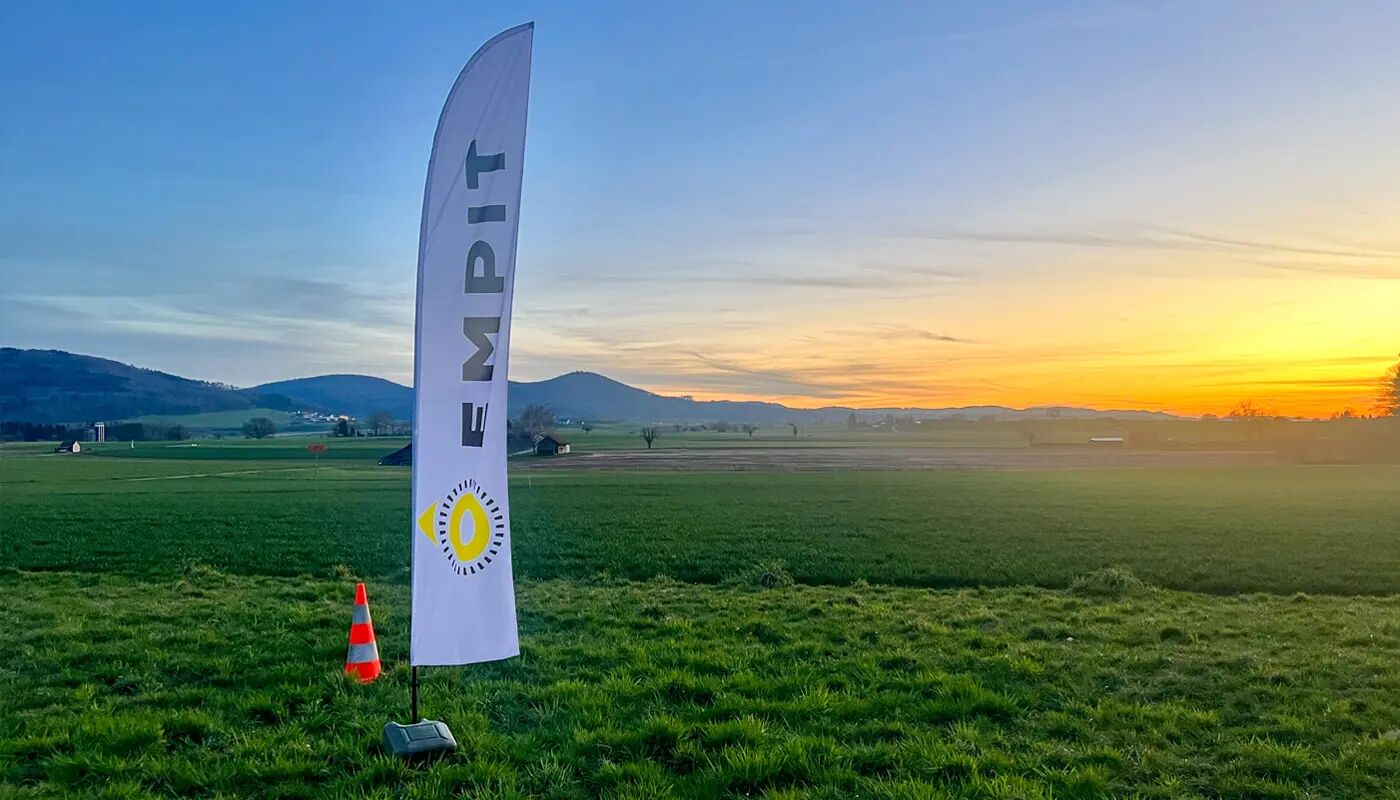 A scenic countryside landscape at sunset featuring an EMPIT branded flag. The flag is placed in an open green field, with rolling hills in the background.