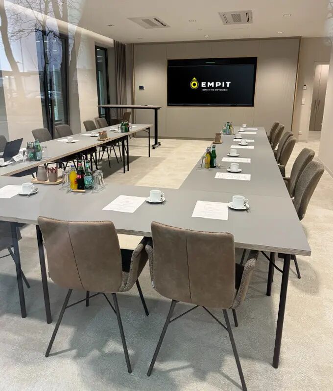 A modern U-shaped conference setup at the Lingen department with grey tables, upholstered chairs, coffee cups, notepads, and refreshments, lit by large windows and featuring a wall-mounted display with the EMPIT logo at the front